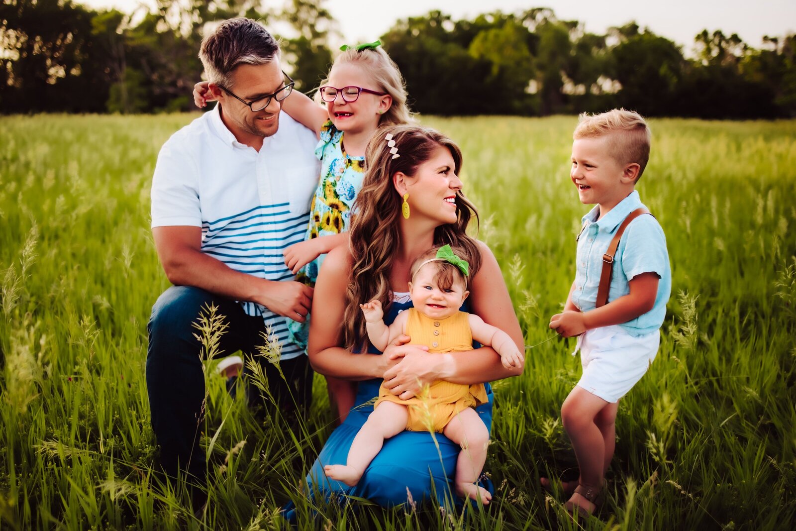 A family of 5, mother, father, and their three children all smiling and laughing with each other in a grass field in Nebraska captured by Infinite Productions
