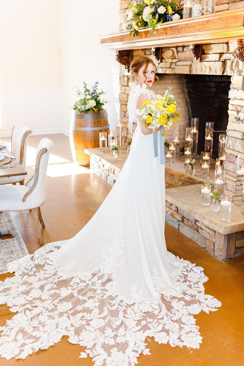 Bride holding a bouquet standing beside the grand stone fireplace at Willow and Stone Events in Mocksville, NC, showcasing elegant wedding decor.