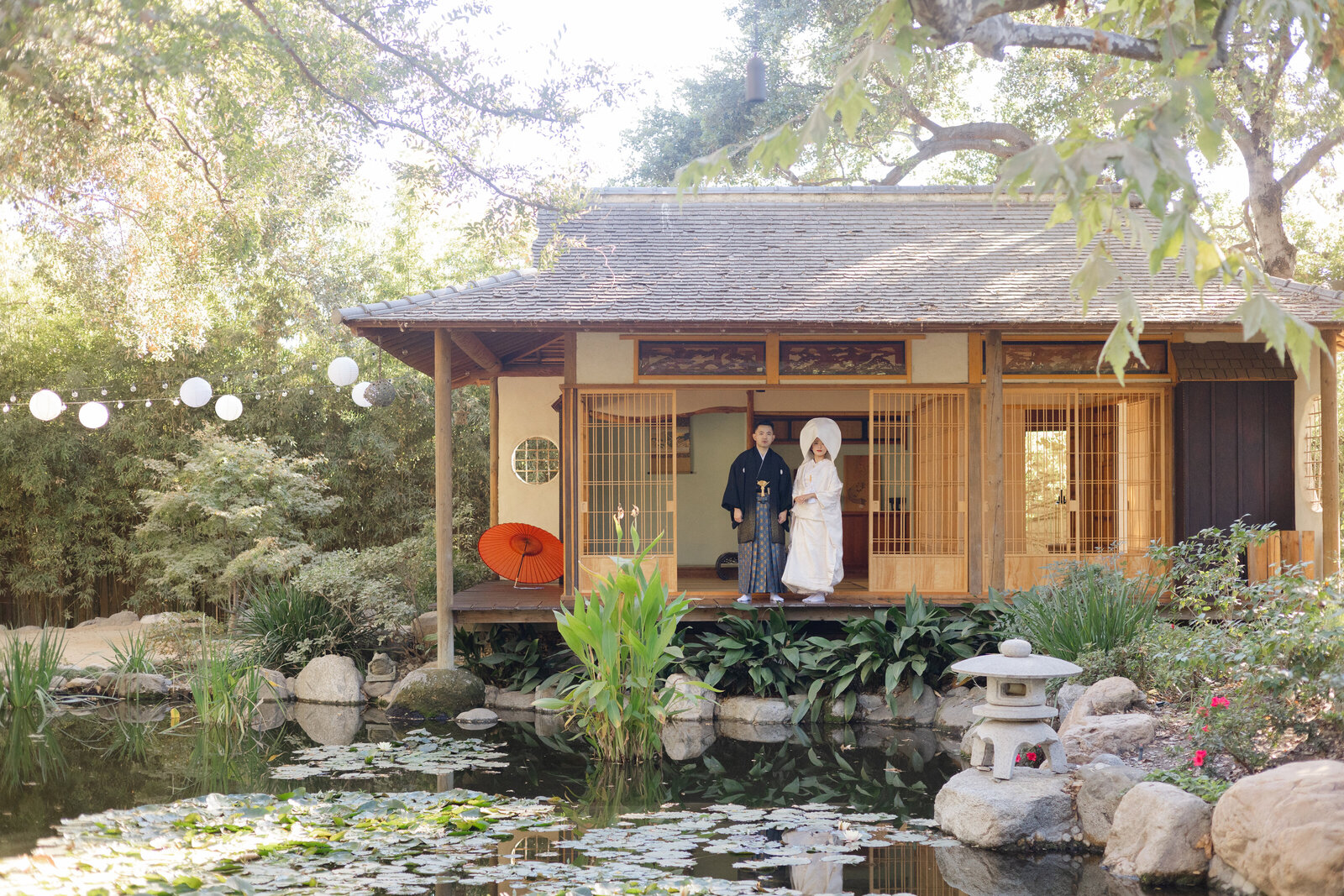 Bride and groom in pure white shiromuku and black montsuki wedding kimono rental, posing beside the pond and tea house at Storrier Stearns Japanese Garden, Los Angeles California | ストーリア・スターンズ日本庭園の池と茶室を背景に白無垢の花嫁と黒紋付袴の新郎が並ぶ。ロサンゼルス カリフォルニアの和装ウェディング着物レンタル。