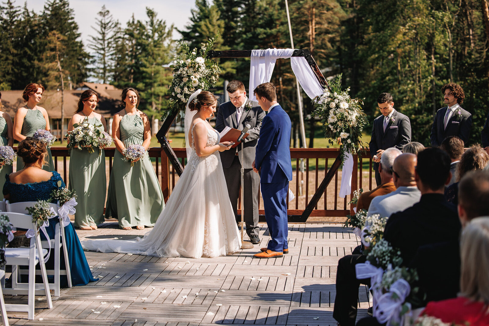 A bride reads her private vows to her groom, standing in front of a wooden arch decorated with white fabric and florals, surrounded by their bridal party and family