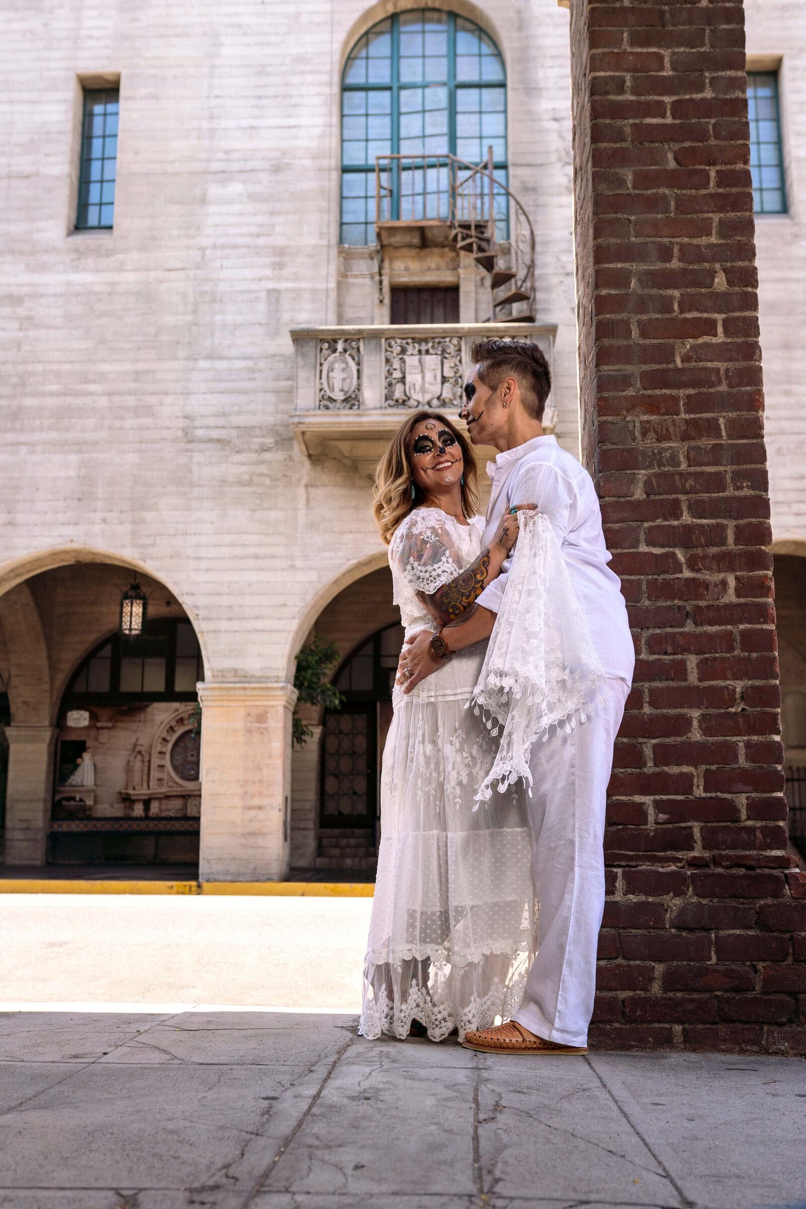 Shorter woman in white dress and painted face hugging fiancee in white  in front of mission inn balcony.