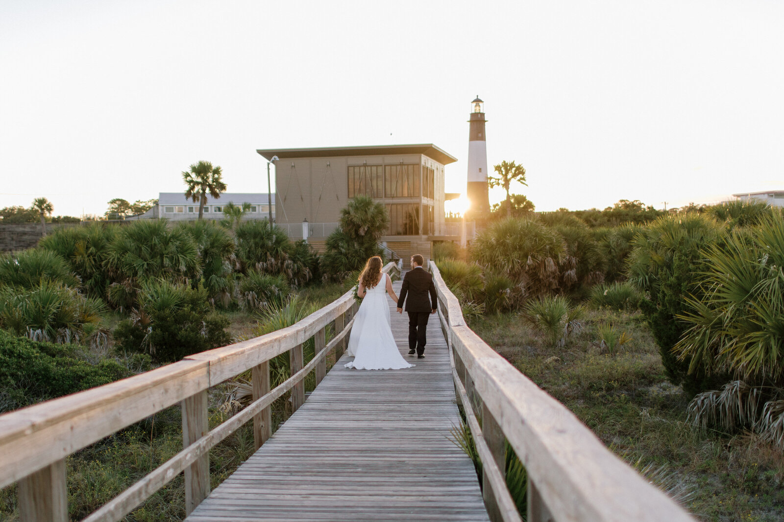 Wedding photography in savannah, ga of bride and groom walking away from camera down beach board walk holding hands at sunset toward a lighthouse with sunset peaking around the side.