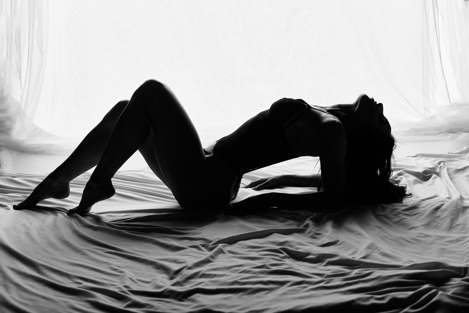 A gorgeous black and white silhouette of a  woman in lingerie laying in bed sheets.