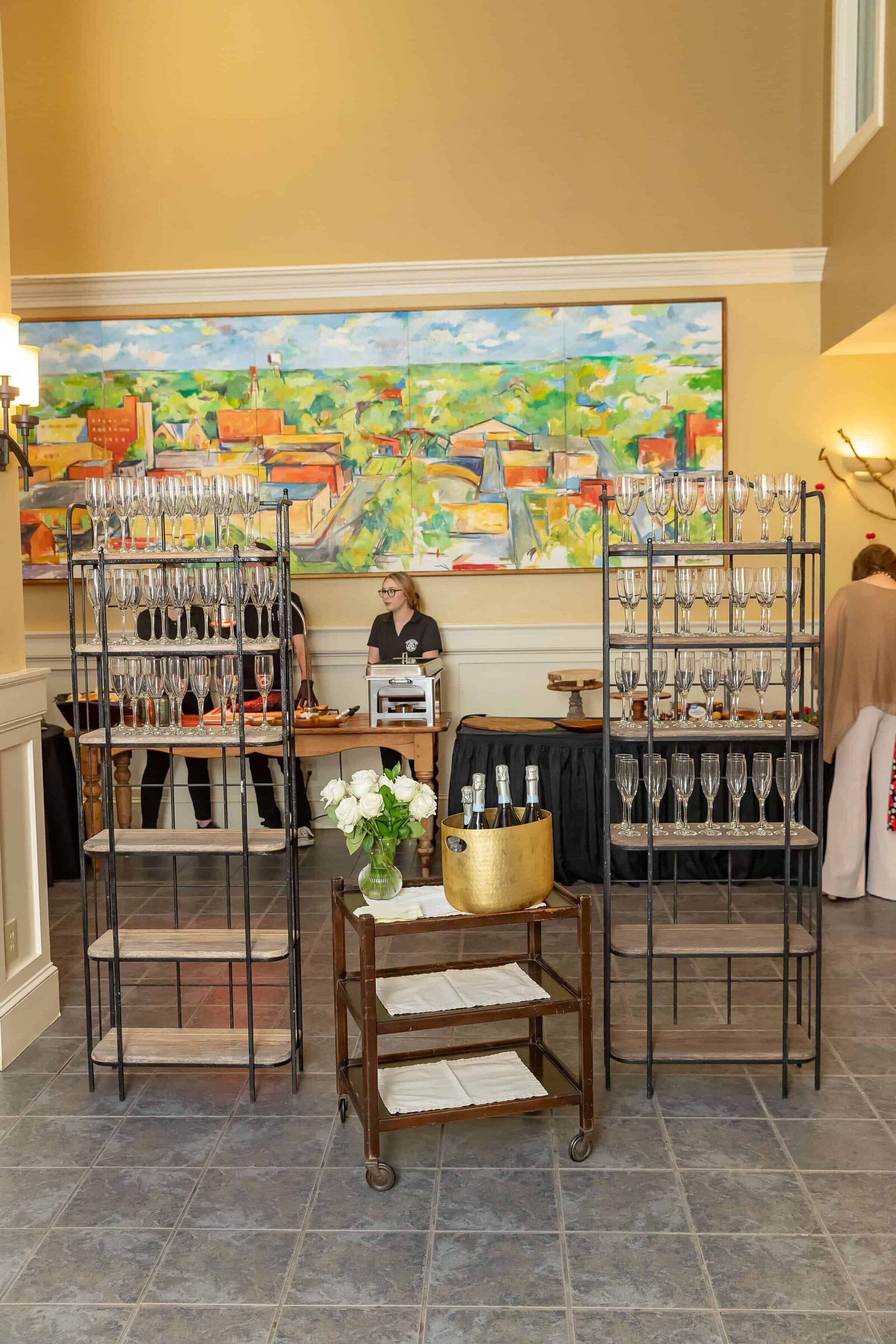 A drink station with racks of empty wine glasses, a cart with a gold ice bucket and bottles, and a person standing behind a table. A colorful painting hangs on the wall in the background.