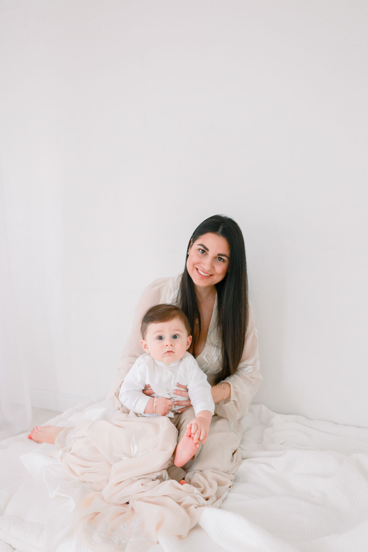 Bei einem Fotoshooting mit Baby im Fotostudio Bielefeld für Babyfotografie strahlt eine Mama mit langen dunklen Haaren und einem langen zarten Kleid vor einem hellen Hintergrund mit ihrem überrascht drein schauendem Baby auf dem Schoß.