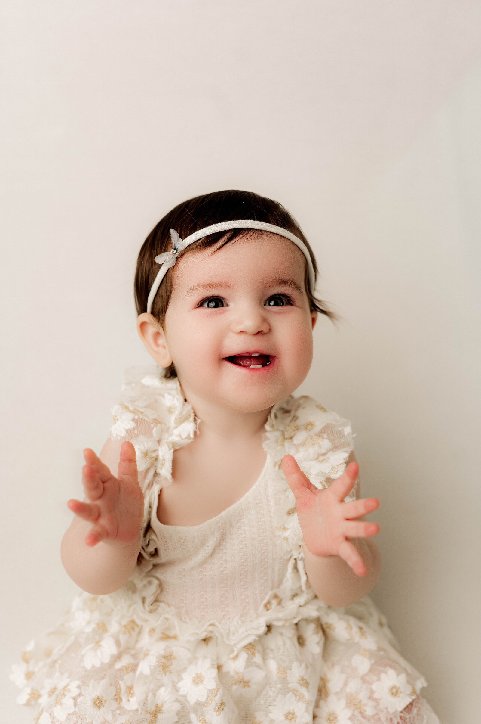 One year old baby girl sitting in a white flower dress clapping her hands