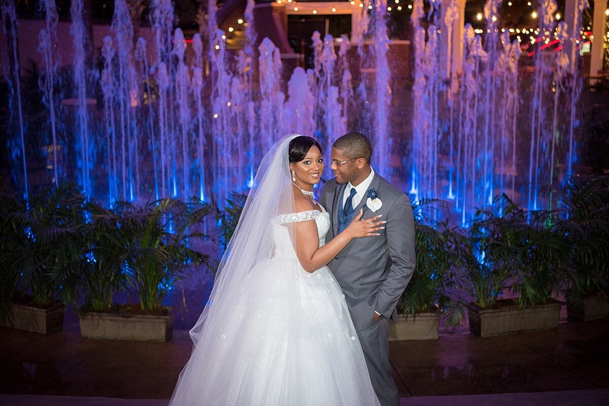 The bride, wearing a full tulle wedding gown and veil embraces the groom wearing a gray suit as the fountain at Opryland Hotel dances behind them with purple uplighting