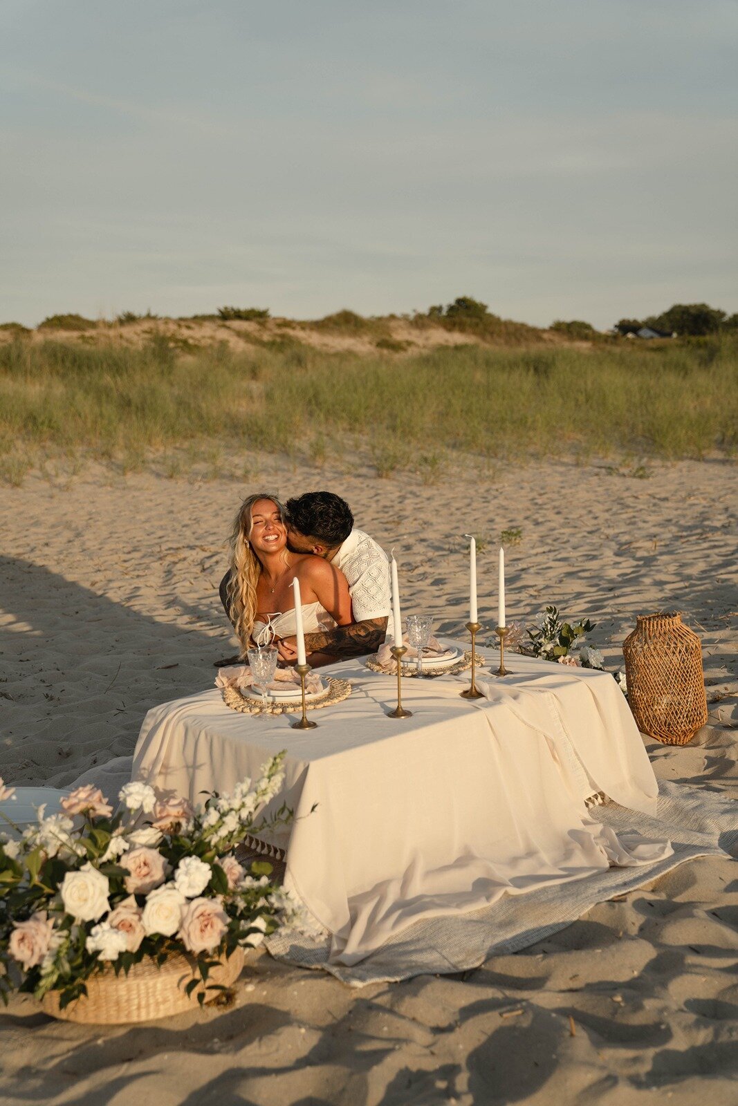 Couple embracing at a romantic beach picnic with elegant table settings, candles, and floral arrangements, styled by Yours Truly, a wedding planner and designer in New Jersey.