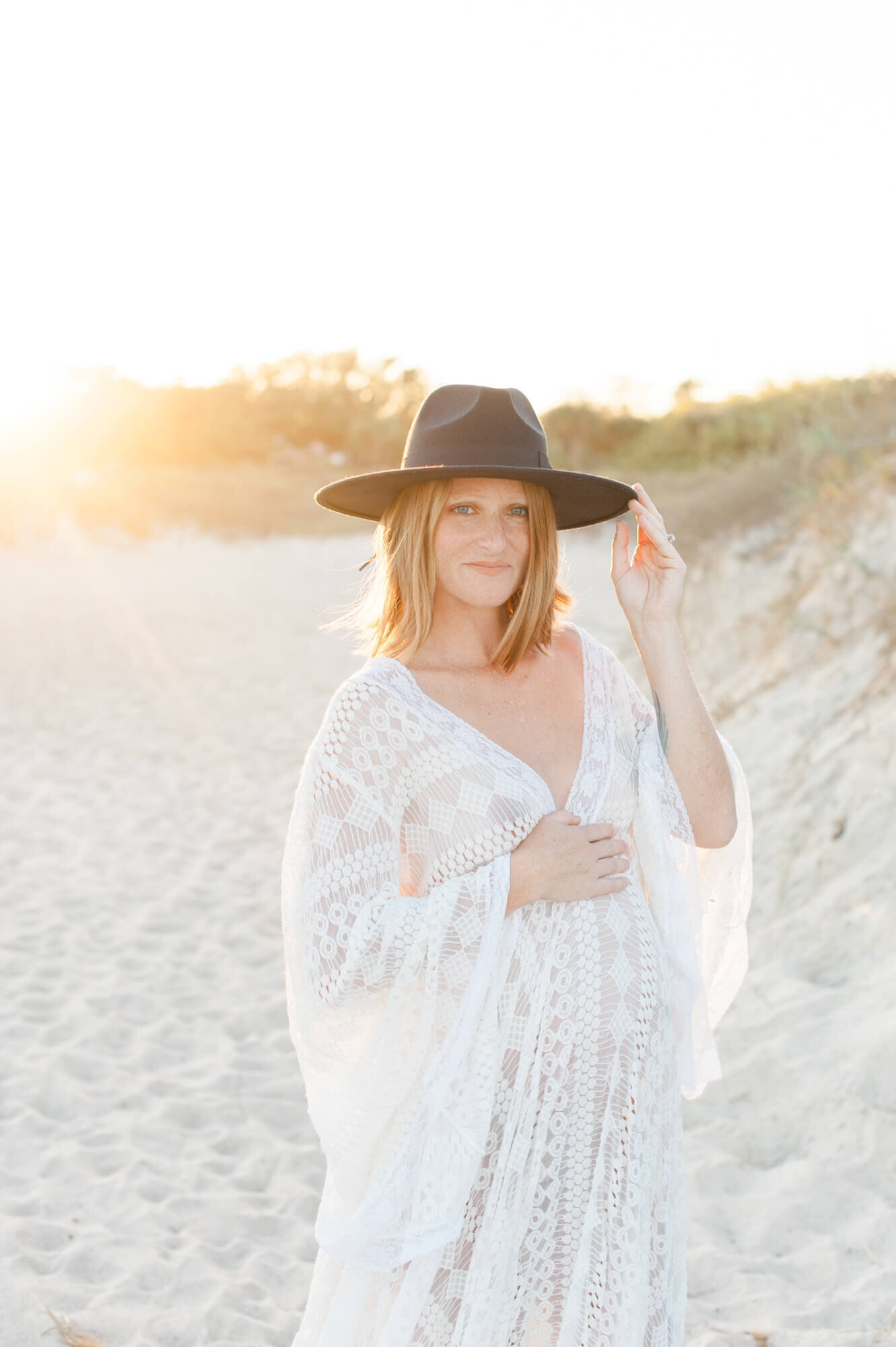 Expectant mother holds her wide brim hat and places her hand on her belly while looking into the camera at sunset