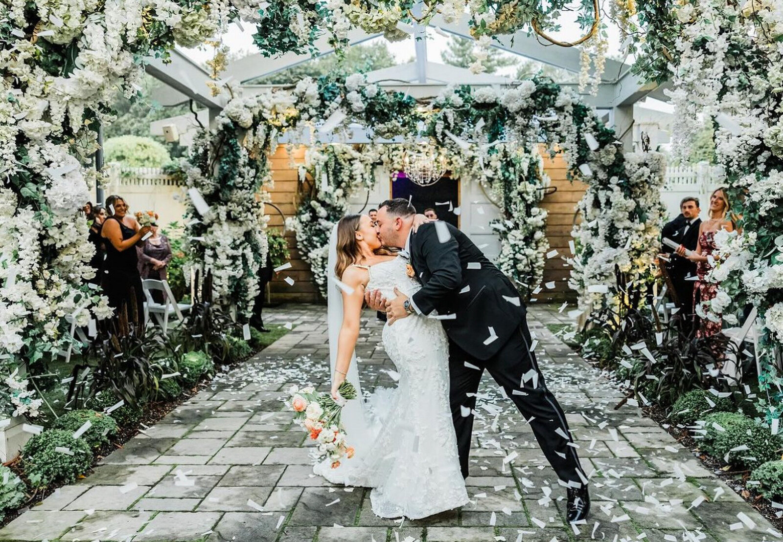 newlywed couple kissing. The bride holds a bouquet of pastel-colored flowers and groom dips the bride under a floral archway adorned with lush greenery and white flowers