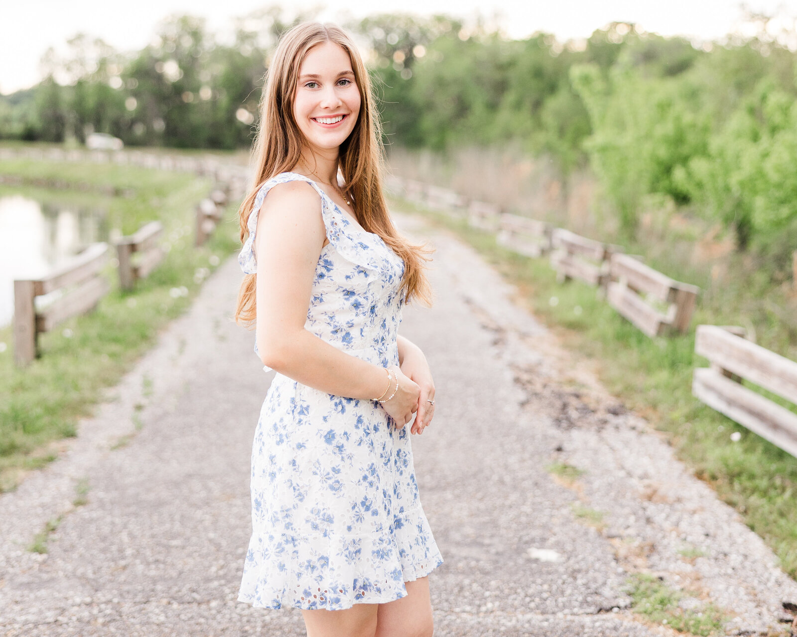 Sophie standing on a lakeside trail, wearing a blue floral dress, during her senior portrait session with Abby Bell Photography, Fine Art Portrait Photographer based in Alabama.