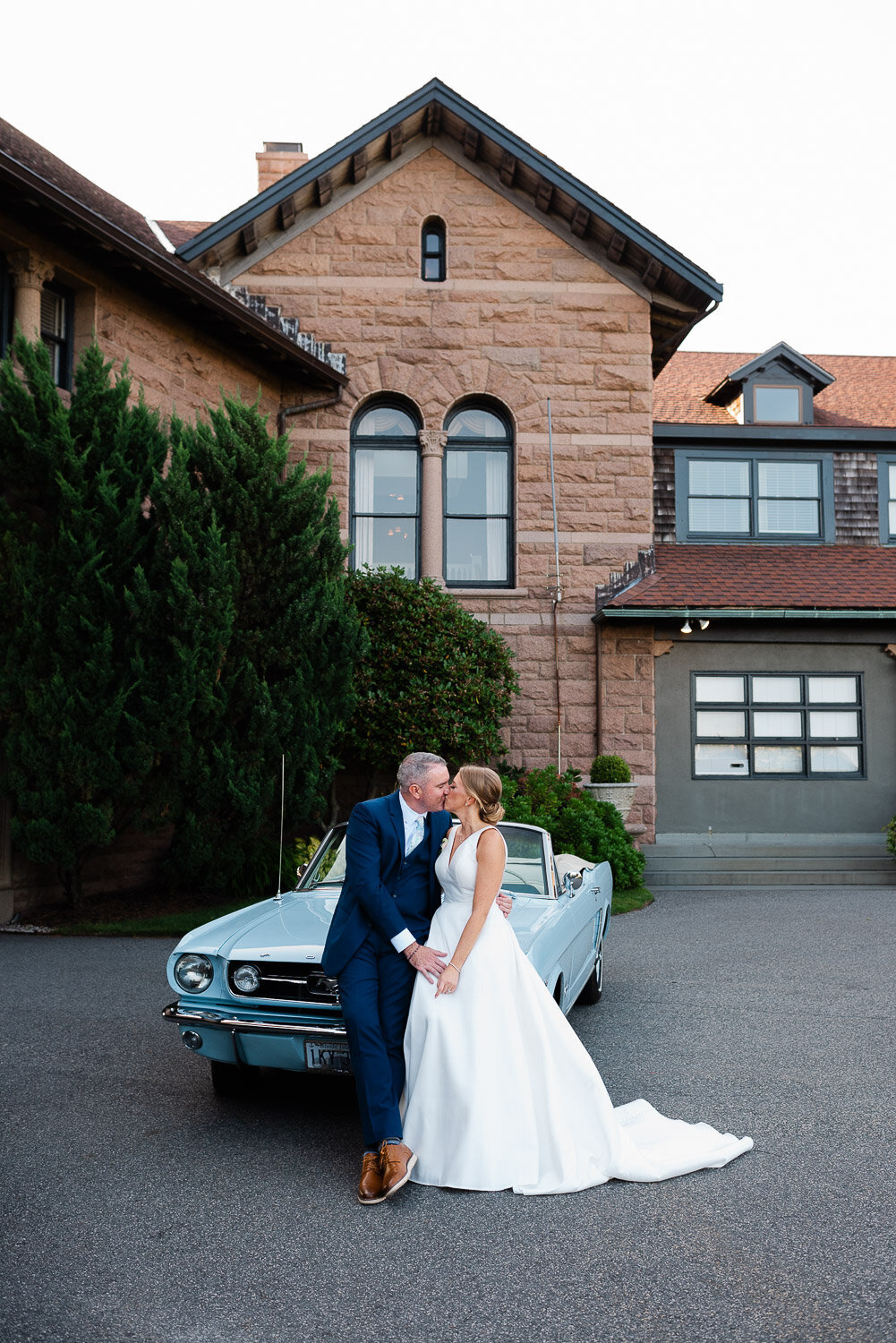 bride and groom kiss on hood of blue car in front of Oceancliff