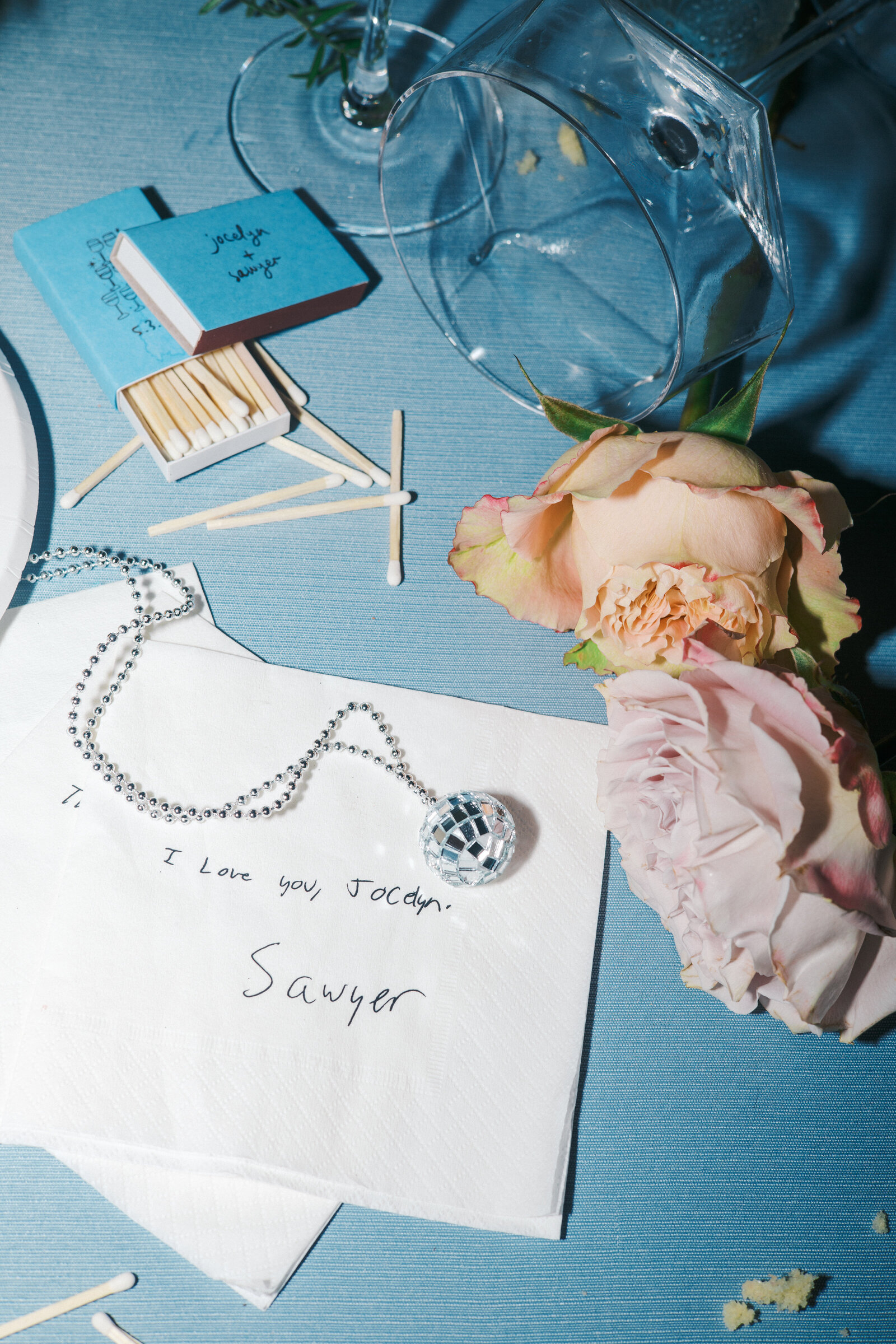 Night time wedding reception flat lay of handwritten note to the. Bride on a cocktail napkin from the groom. Matches and disco ball necklace