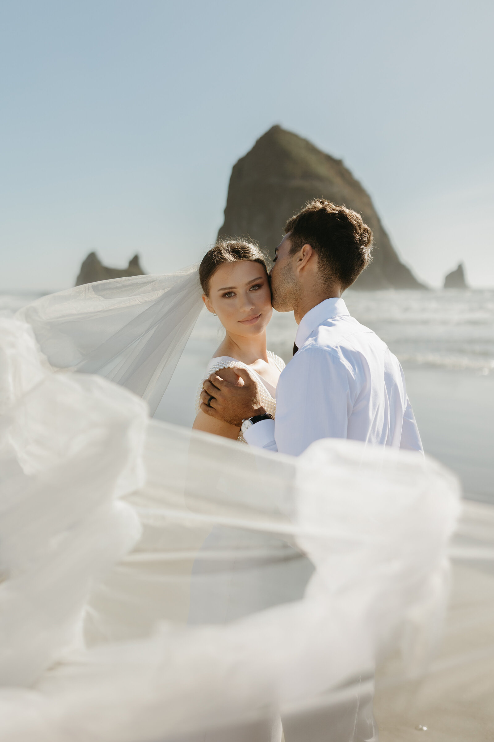 A bride looks into the camera while her groom kisses her with a dramatic veil effect from the windy Oregon beach. 