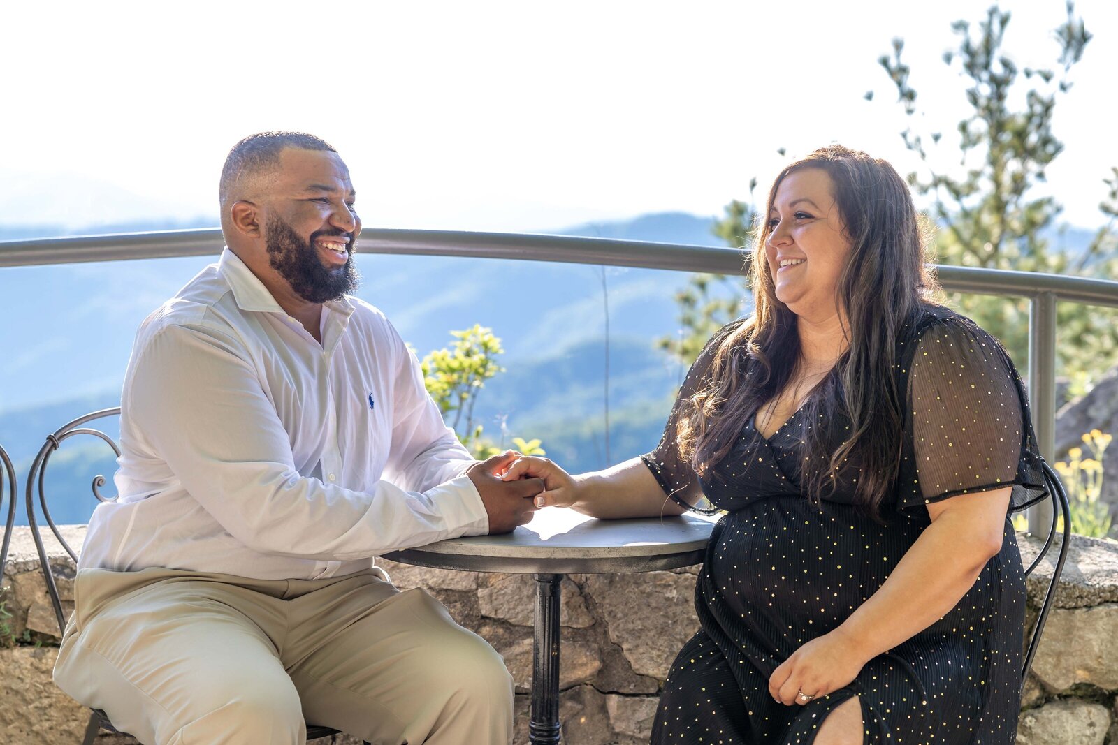 A smiling couple enjoys a serene moment at an outdoor patio with stunning mountain views in the background. The man, dressed in a crisp white shirt, holds hands with the woman, who wears a black polka-dot dress. This cheerful scene captures the natural beauty of North Carolina engagement photography. 