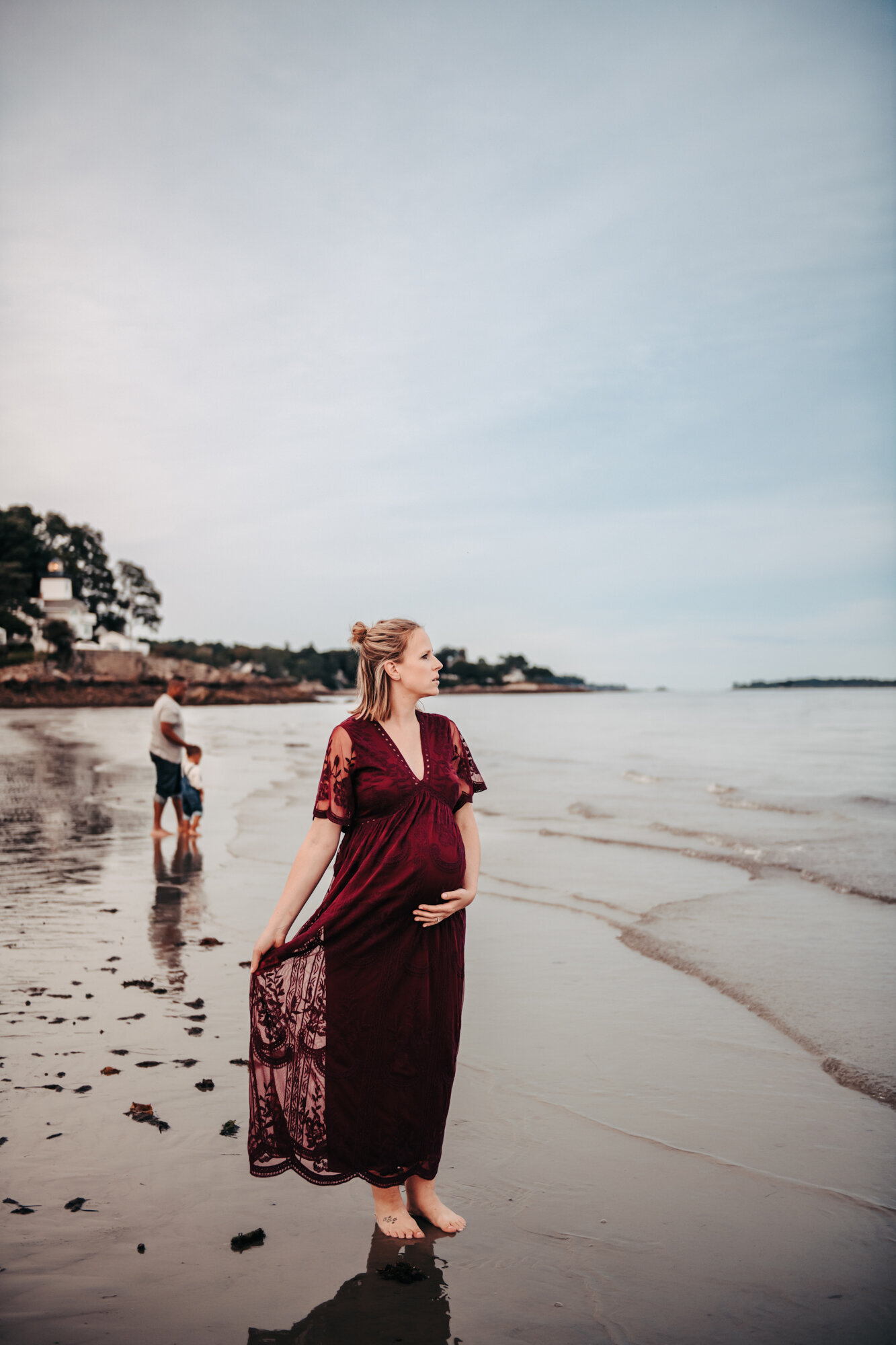 Maternity Photographer, A pregnant mom in a  dress stands barefoot on the beach while a man and little boy play behind her.