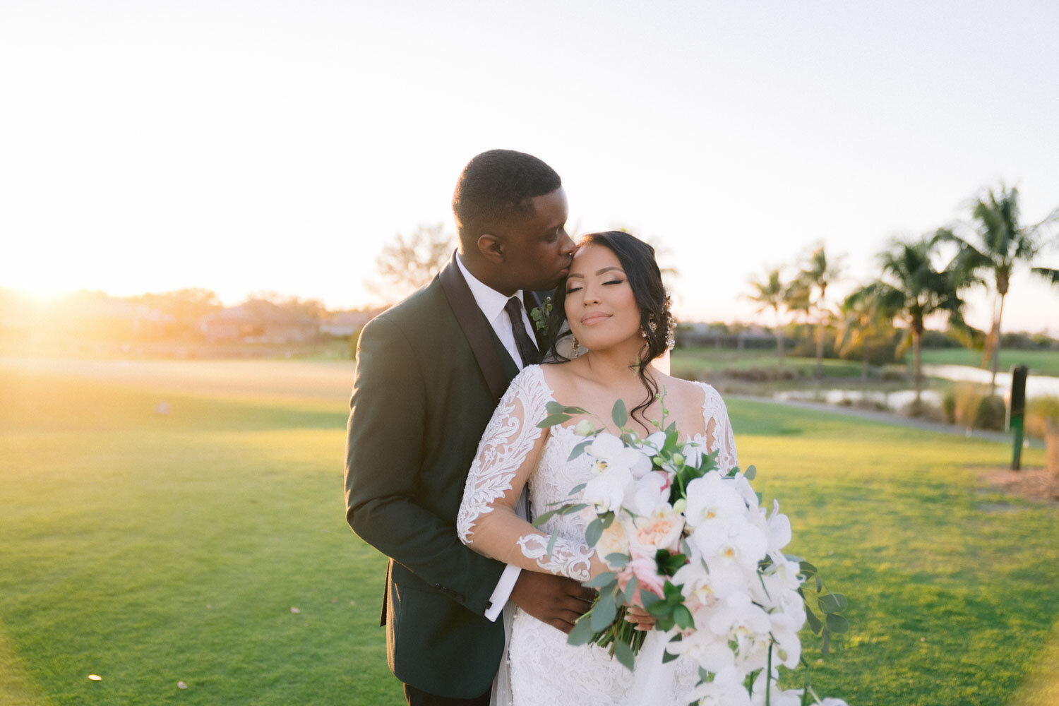 Groom kissing bride's head as they embrace in a sunlit outdoor setting