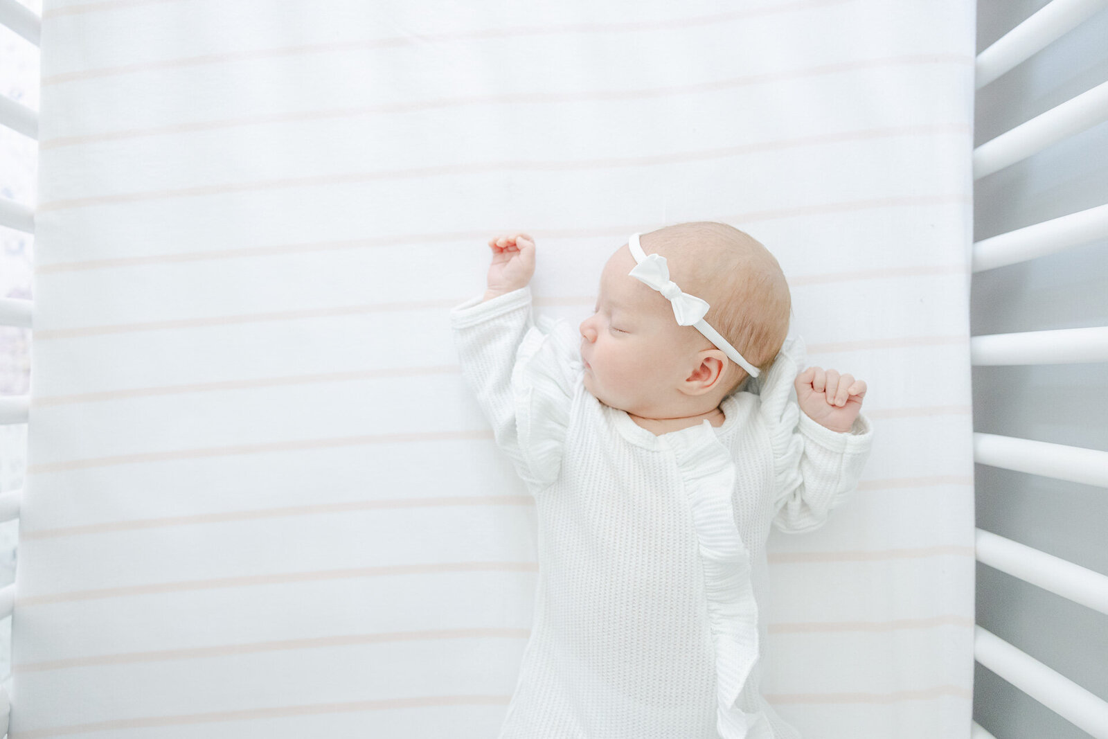 Newborn baby girl in a white onesie and bow headband peacefully sleeping on a soft striped blanket during an in-home lifestyle session with Connecticut newborn photographer Nicole Neira.