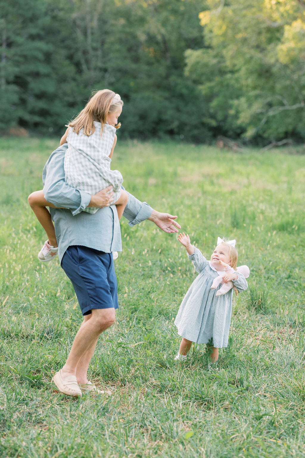 Little girl reaches for dad in a field while dad holds other little girl