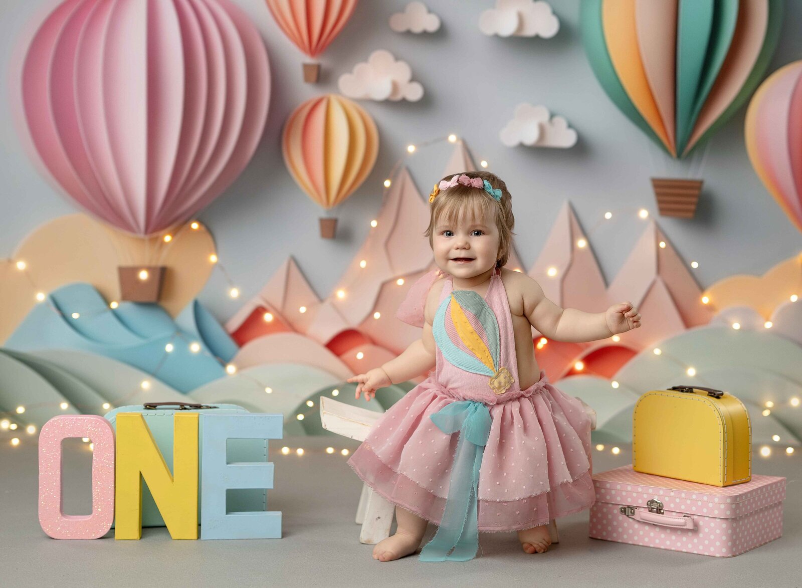 Baby girl in pink dress, sitting on stool in front of hot air balloon backdrop for 1st birthday photos in Atlanta.