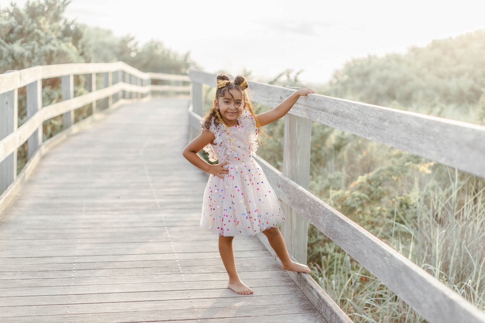 Young girl in a colorful birthday dress posing on a wooden boardwalk with the Fire Island Lighthouse in the background.