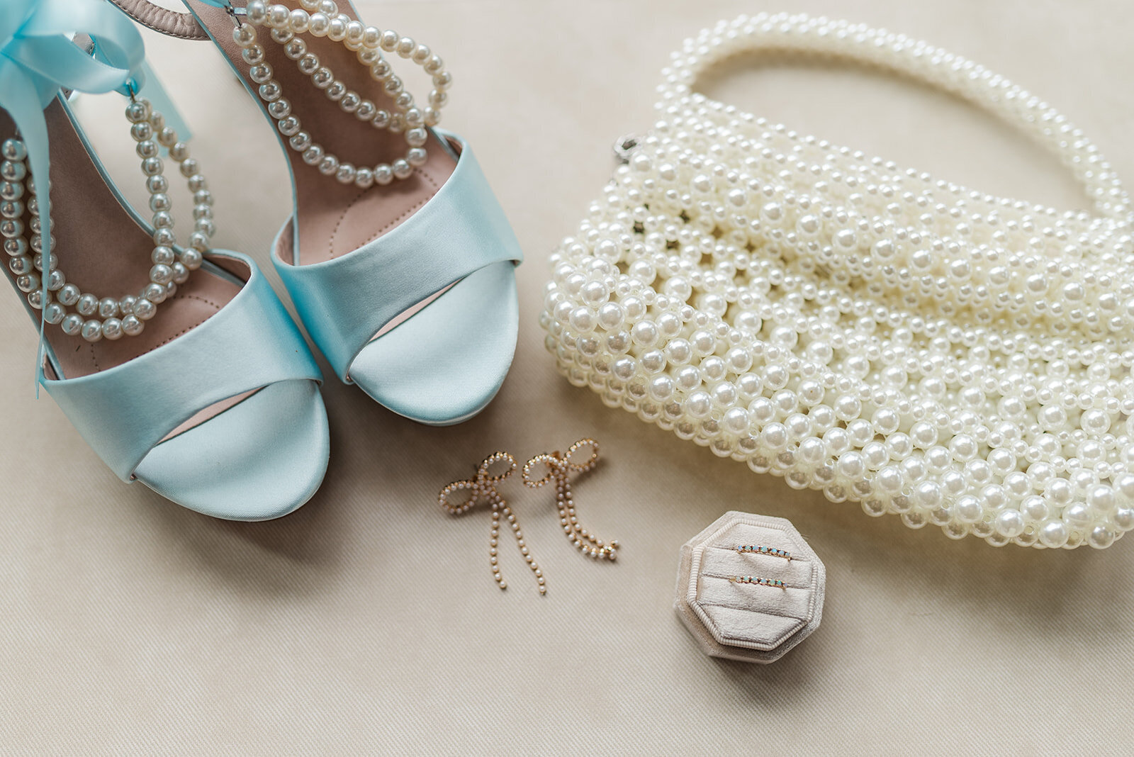 flat lay of blue wedding heels, pearl clutch, bow sparkle earrings, and ring box with wedding rings on a off-white canvas background