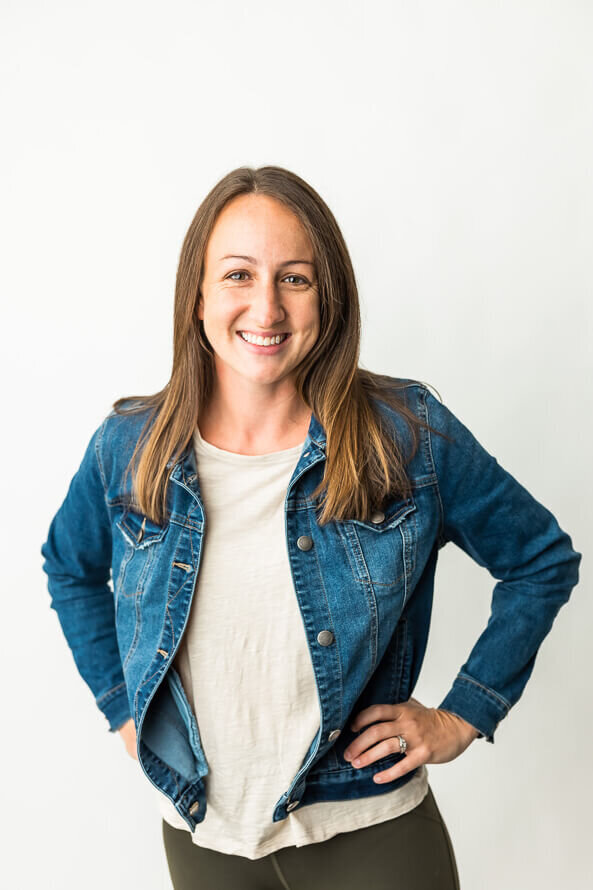 Girl in jean jacket puts hands on hips for her headshot in the white photo studio.