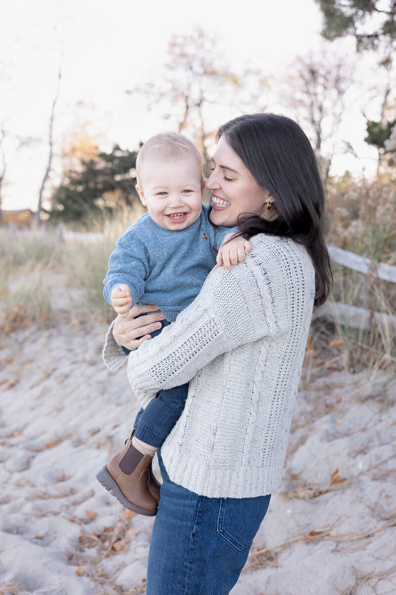 Mother holding her smiling baby boy during a cozy fall family session on the beach in Westchester County, NY. Captured by Nicole, a lifestyle family photographer serving Connecticut and Westchester with a natural, joy-filled approach.