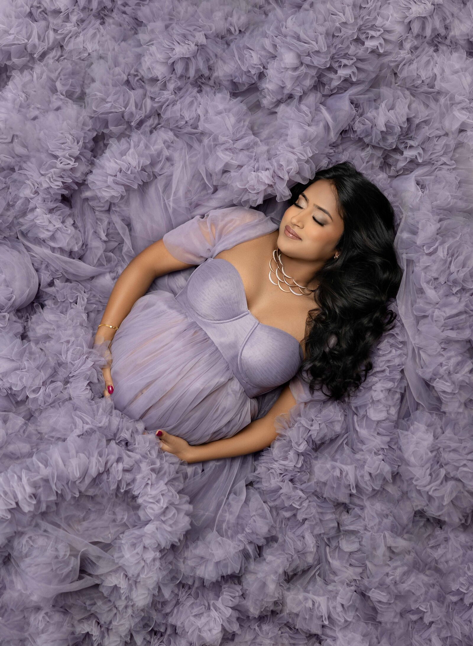 Pregnant women laying in lavender tulle and holding her belly with eyes closed.