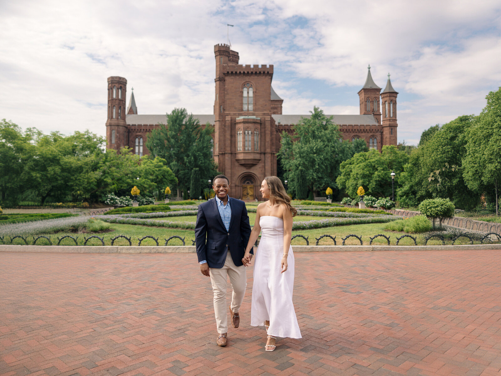A couple walks hand in hand on a brick path in front of a historic building with towers. The man wears a blue blazer and beige pants, and the woman wears a white dress. Lush greenery surrounds them under a partly cloudy sky.
