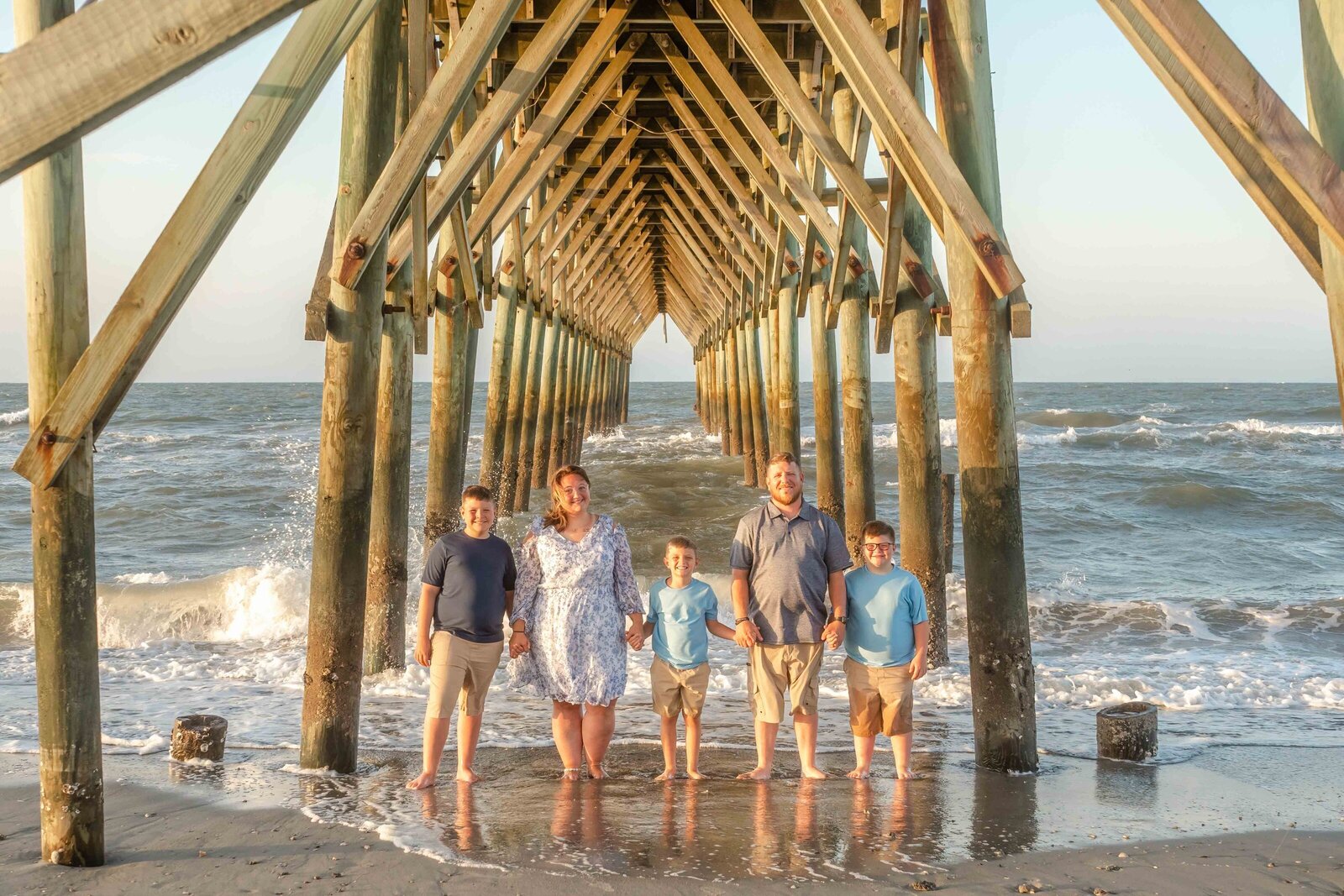 A family of five holds hands in the surf beneath a pier, dressed in blue and khaki outfits. The ocean waves and wooden structure provide a dramatic backdrop. Perfect for artistic family photography in Wilmington, NC, highlighting dynamic and memorable moments.