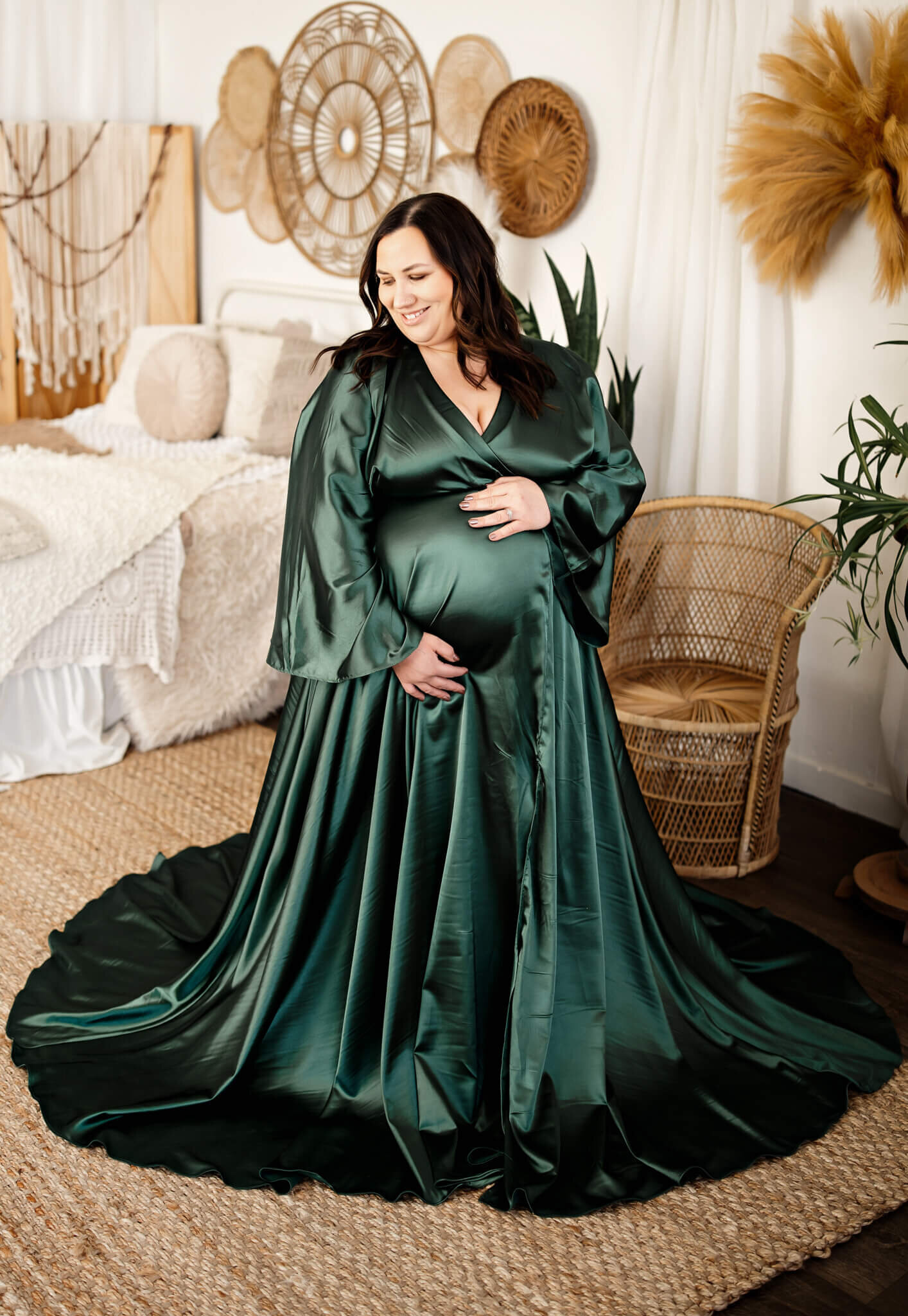 A pregnant woman in a long, flowing emerald green gown poses indoors, cradling her belly and smiling. The cozy room, perfect for maternity portraits, features woven wall art, pampas grass, plants, and a welcoming bed.