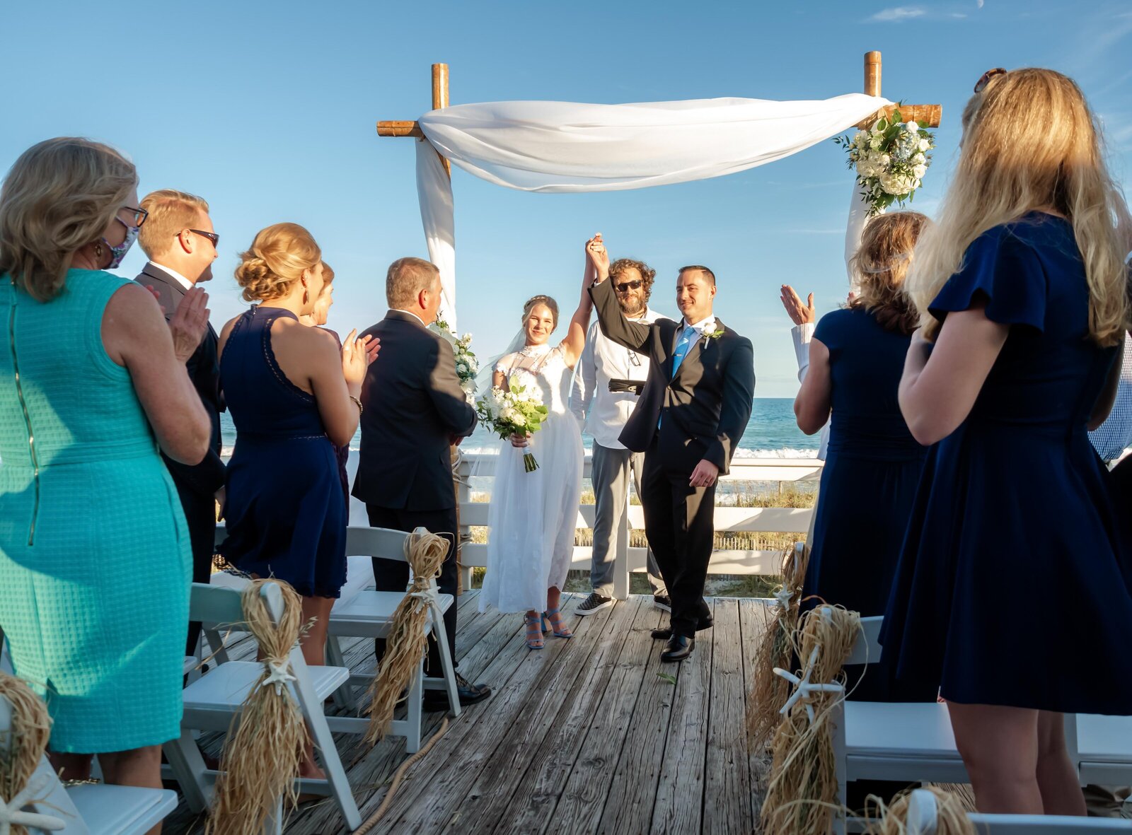 The bride and groom raise their hands in celebration while walking down the aisle at a beach ceremony, surrounded by family and friends, captured at a Topsail Beach, NC wedding, blending joy and coastal charm.