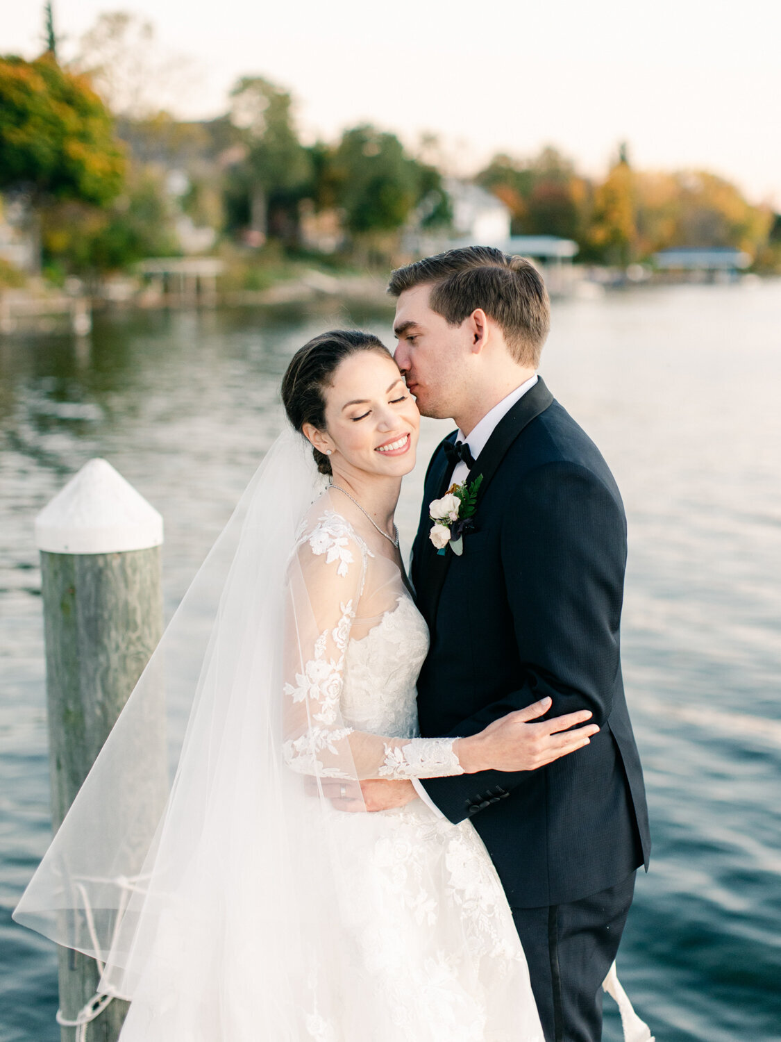A groom gives his bride a kiss on a dock with the Finger Lakes behind them.