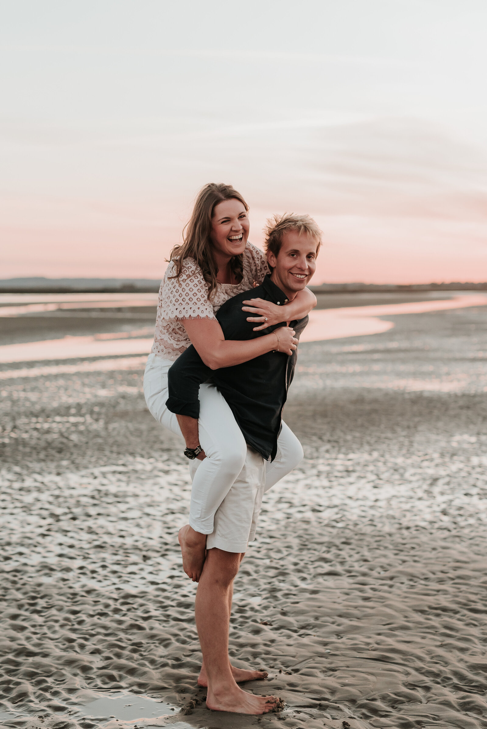 Couple piggyback smiling on a sandy beach as the sun sets at Camber Sands Beach