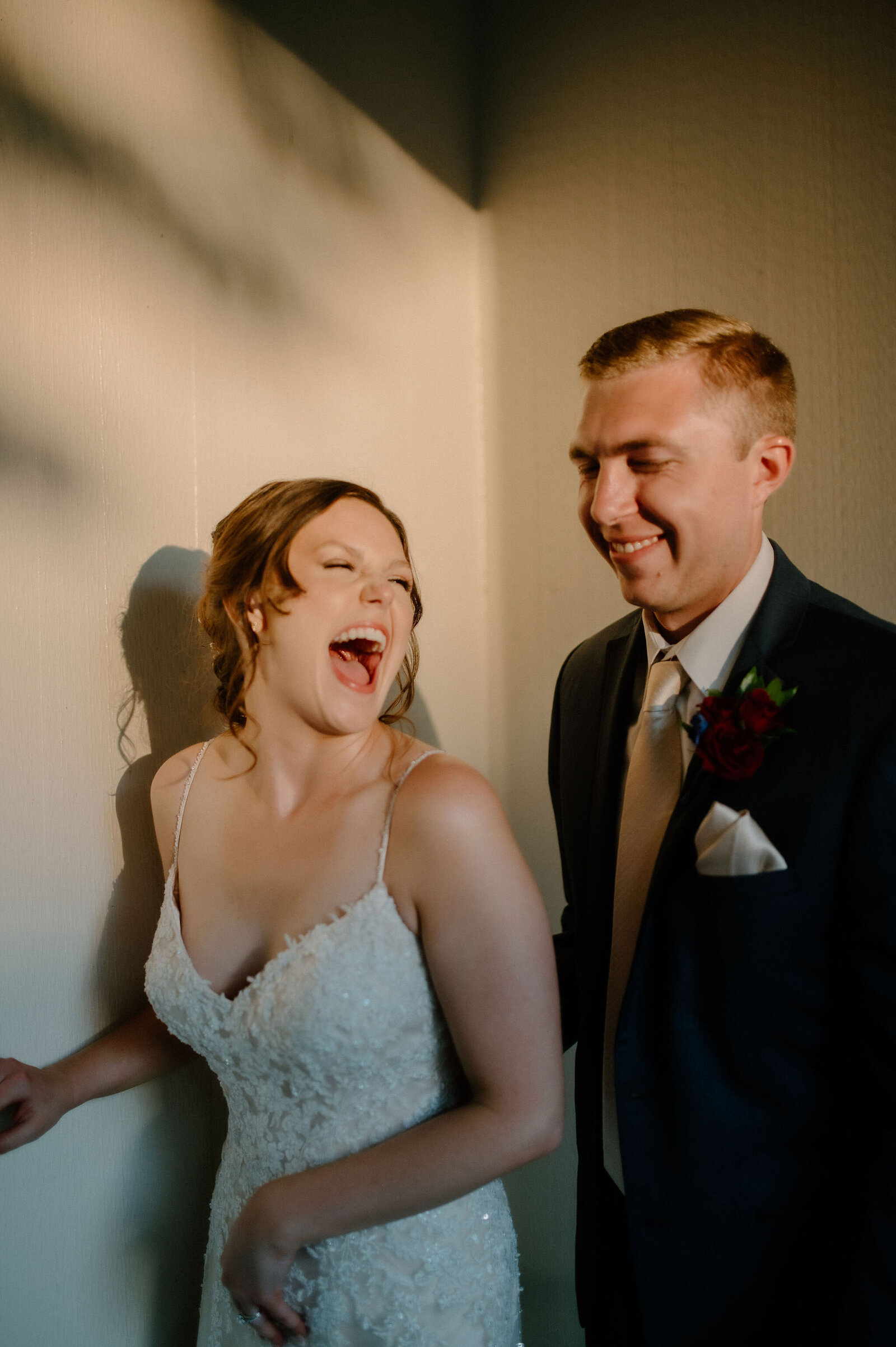 bride and groom standing in corner of room with natural light laughing wedding portrait