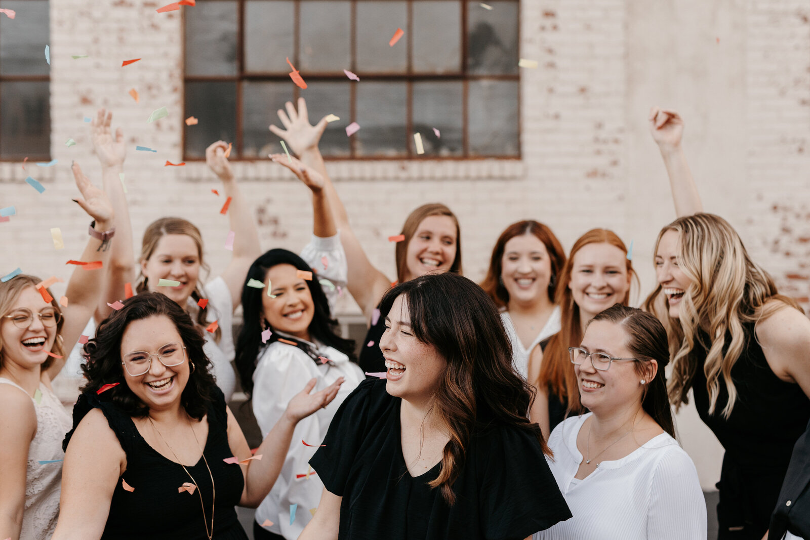 A group of bridesmaids and groomsmen celebrates around a bride and groom as he kisses her cheek at The Hudson event venue in Wichita, Kansas. The bride holds a beautiful bouquet,  the women are wearing different colors of dresses, and the men are waring tuxedos.