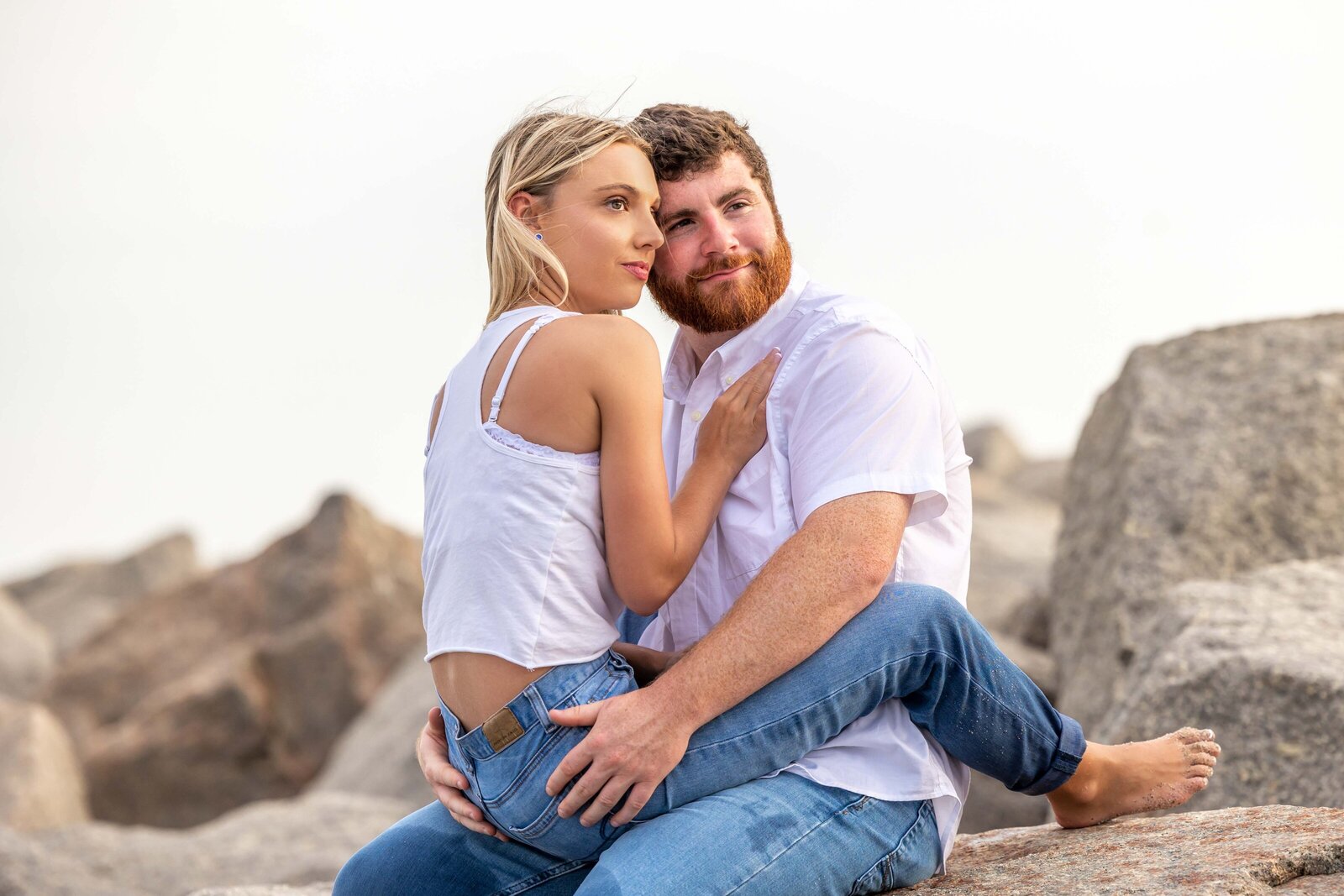 An intimate couple sits closely together on the rocky Fort Fisher beach, with the woman leaning against the man’s chest. Both dressed casually, they share a quiet and tender moment, reflecting the raw and genuine emotions captured in North Carolina wedding photography.