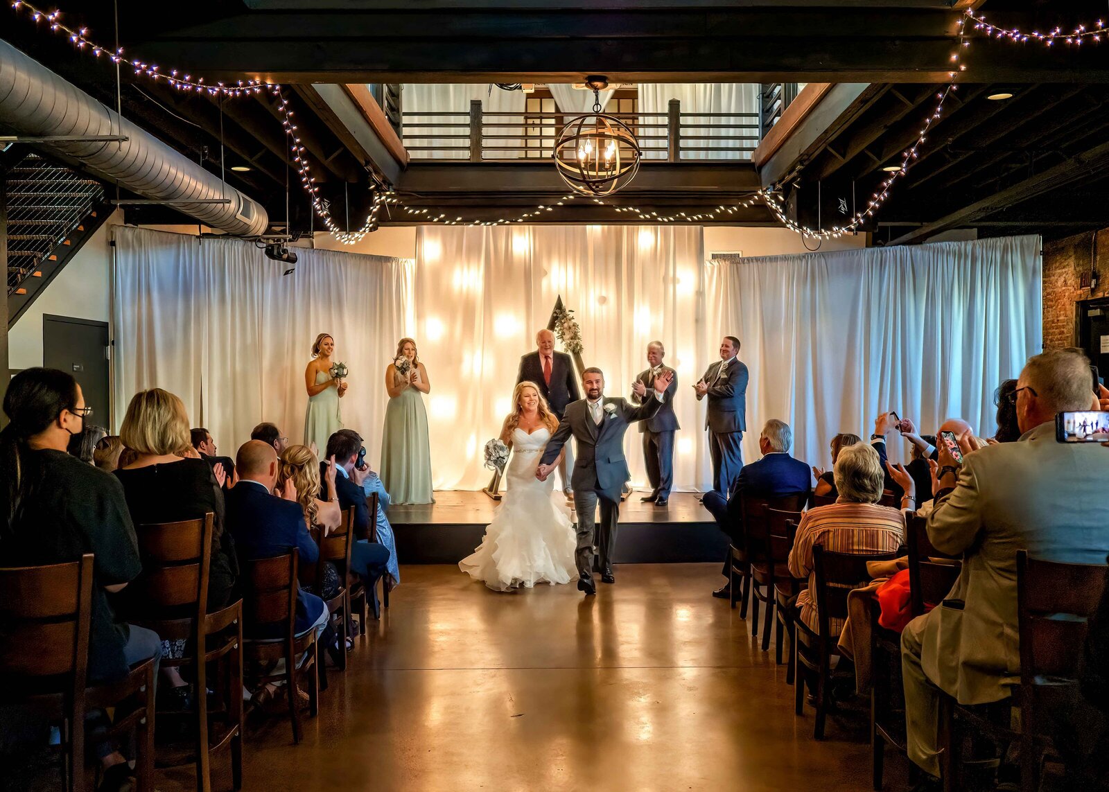 The bride and groom joyfully walk down the aisle after their ceremony, surrounded by twinkling lights and cheering guests, captured during a Wilmington, NC wedding at a modern indoor venue.