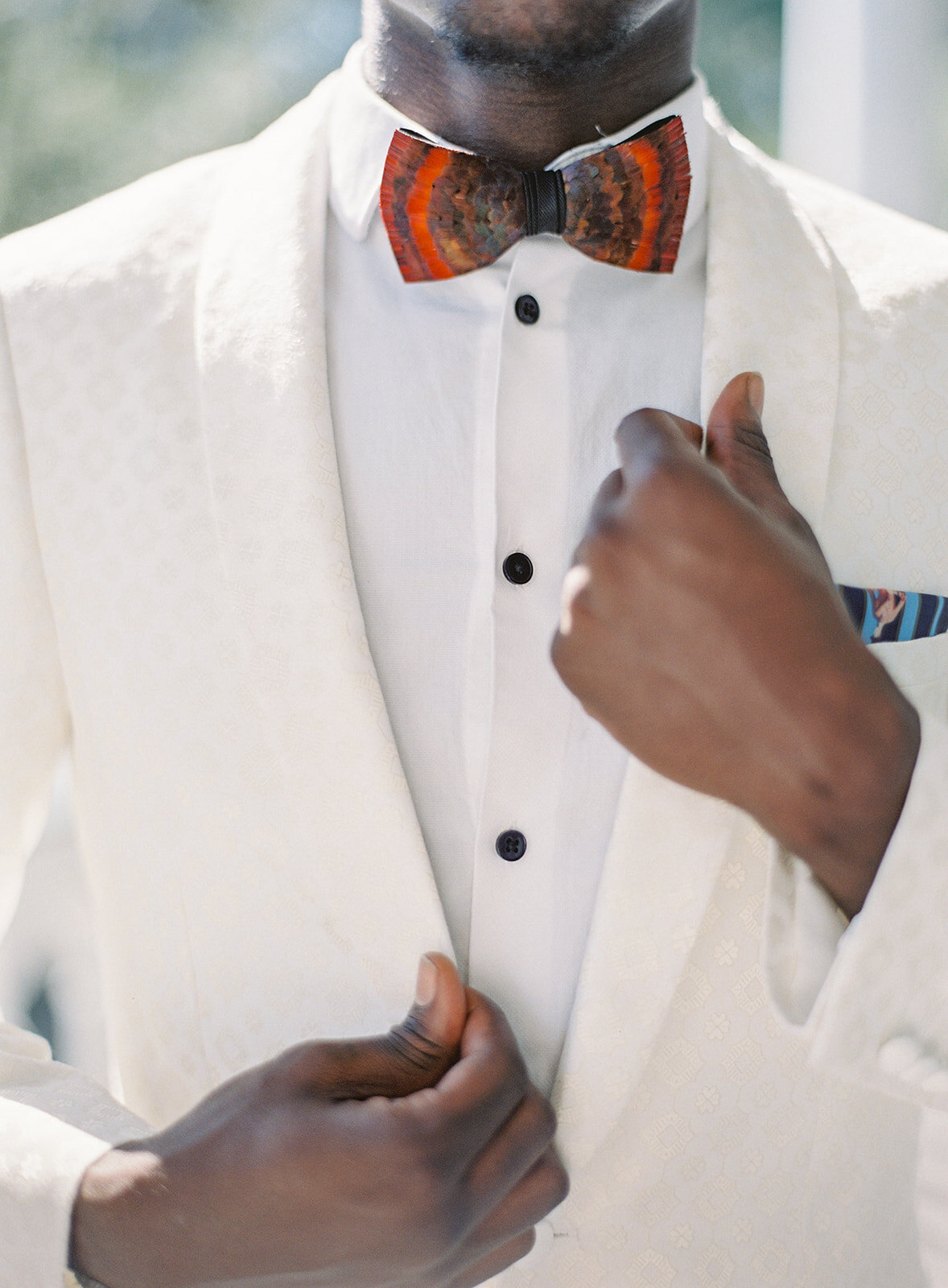 Chest of groom in an ivory and gold detailed tuxedo. He is holding one hand high up on his lapel and the other hand lower on a lapel. He has on a white tuxedo shirt and red pheasant bowtie. Photographed by wedding photographers in Charleston Amy Mulder Photography.