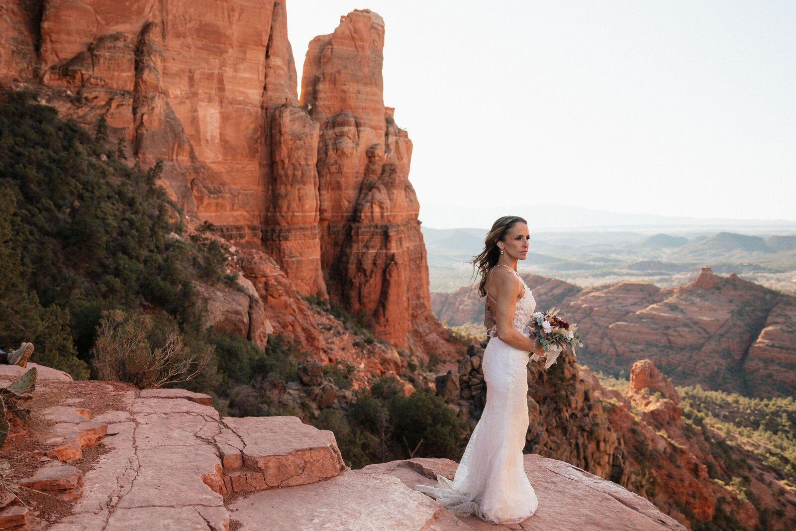 Bride standing on a red rock ledge at Cathedral Rock in Sedona, holding a boho bouquet and wearing a lace gown during her adventurous elopement.