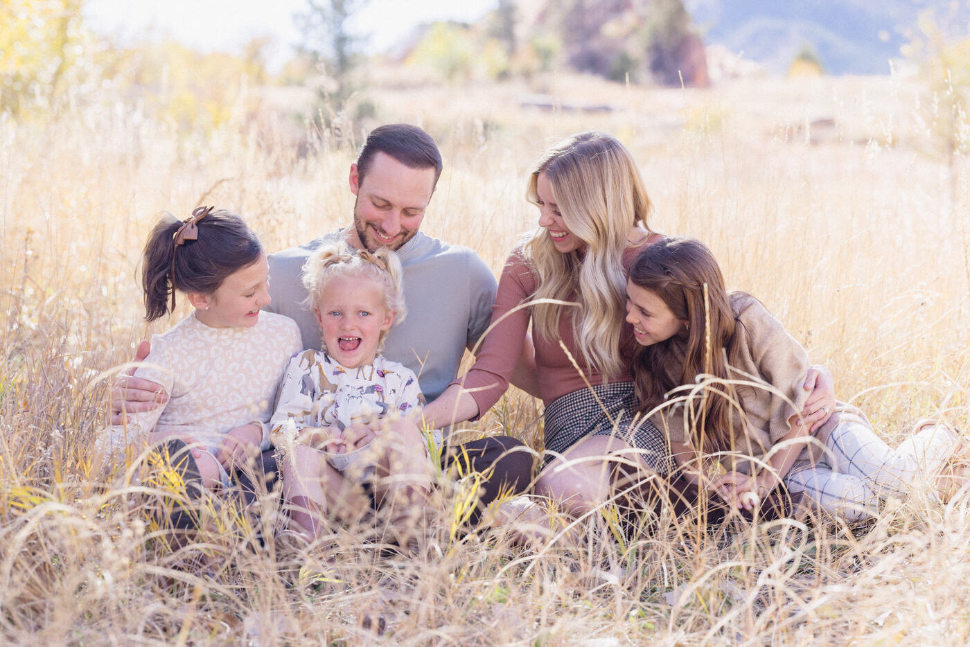 A family of five sits in tall yellow grass looking at each other and smiling.