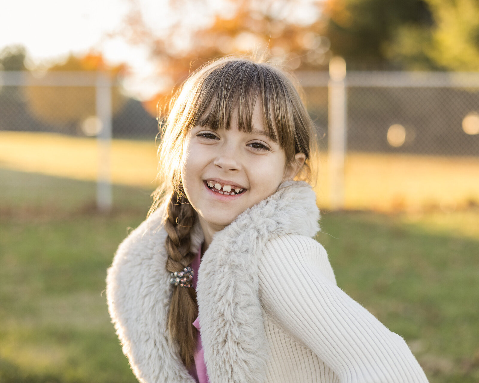 A young girl standing in the grass at an angle, with braided brown hair, a light tan sweater over a pink shirt, smiling at the camera