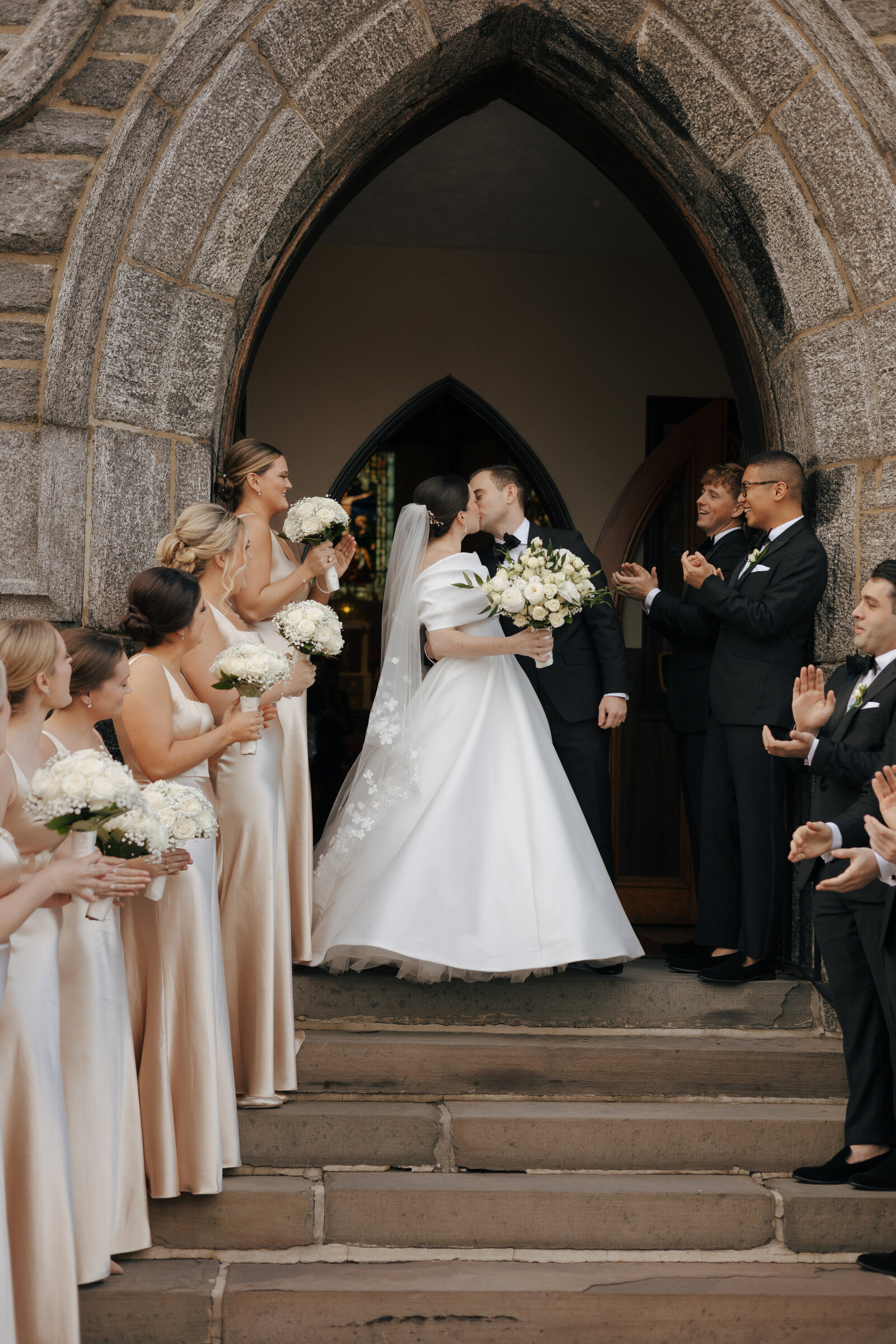 Bride and groom kissing at the outside stairs of their wedding chapel surrounded by their bridesmaids and groomsmen | Emma LeeAnn Photography