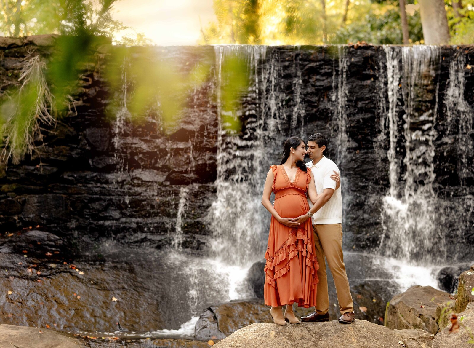 Man and pregnant women standing on a rock by a large waterfall near Atlanta. Both holding baby bump and cuddling.