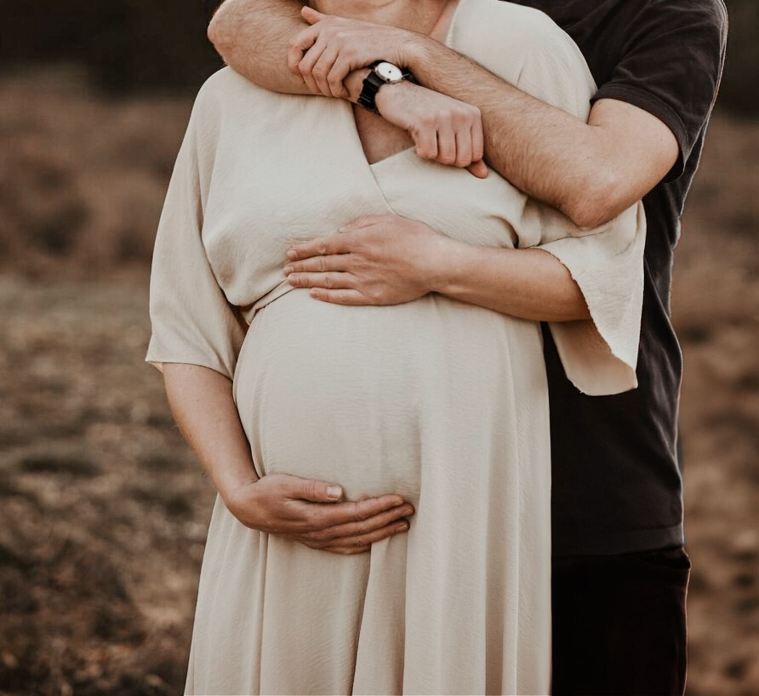 Een close-up van een zwangere vrouw die omhelsd wordt door haar partner, met de focus op hun handen op haar buik