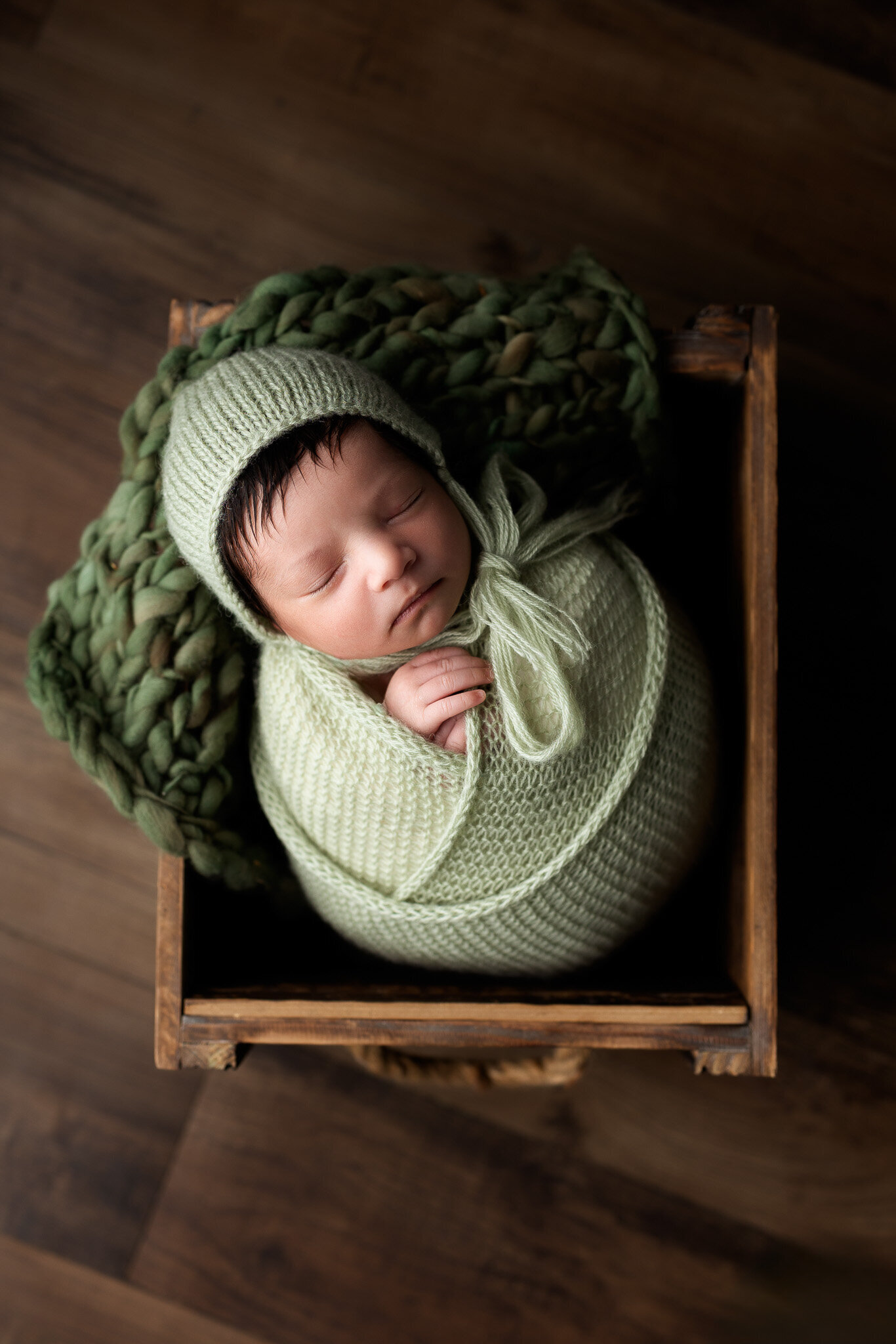 Top down view of a newborn baby boy wrapped in a sage green knit wrap, posed in a rustic wooden crate