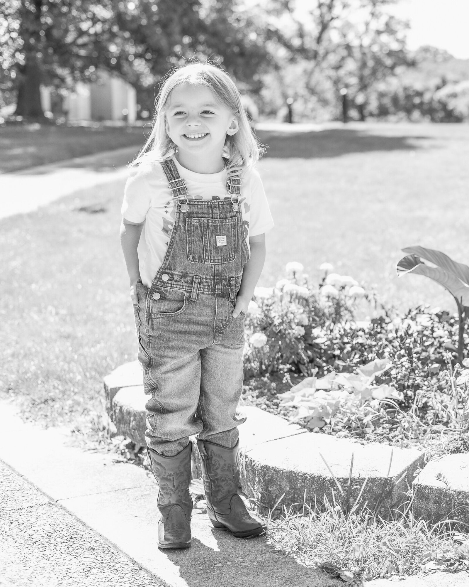 A black and white photo of a young girl in a park wearing jean overalls with each of her hands in a pocket, a white print shirt under the overalls and is smiling off camera.