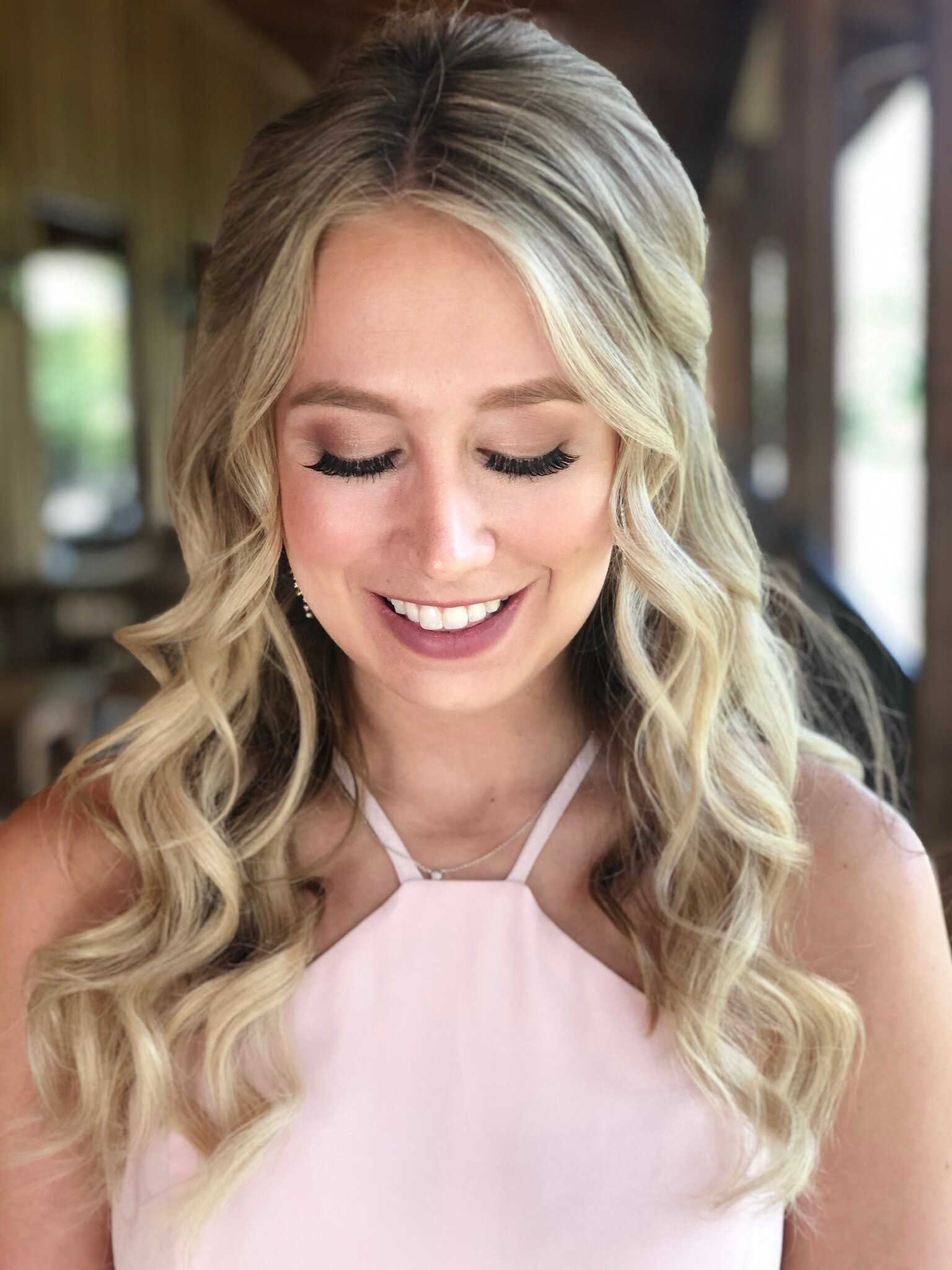 Woman with long wavy blonde hair and a pink top smiles softly while looking down, capturing the serene essence of a bridal beauty experience in Texas.