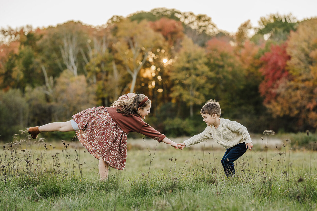 siblings stand on tip toe and reach for each other in a field with autumn colors in boston